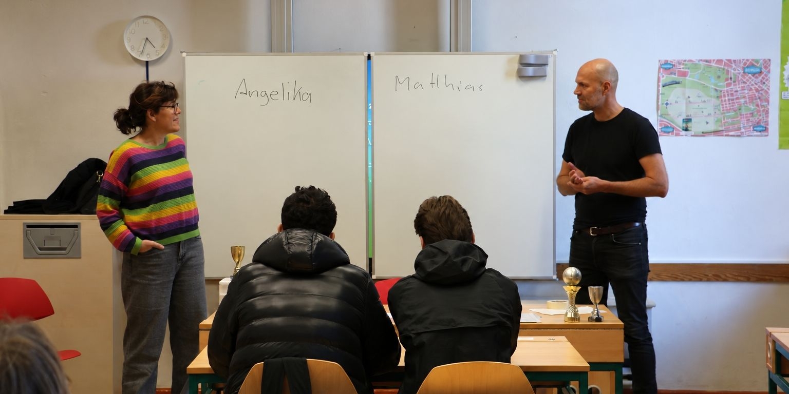 Zwei Teilnehmende sprechen mit Jugendlichen vor einer Tafel im Klassenraum.